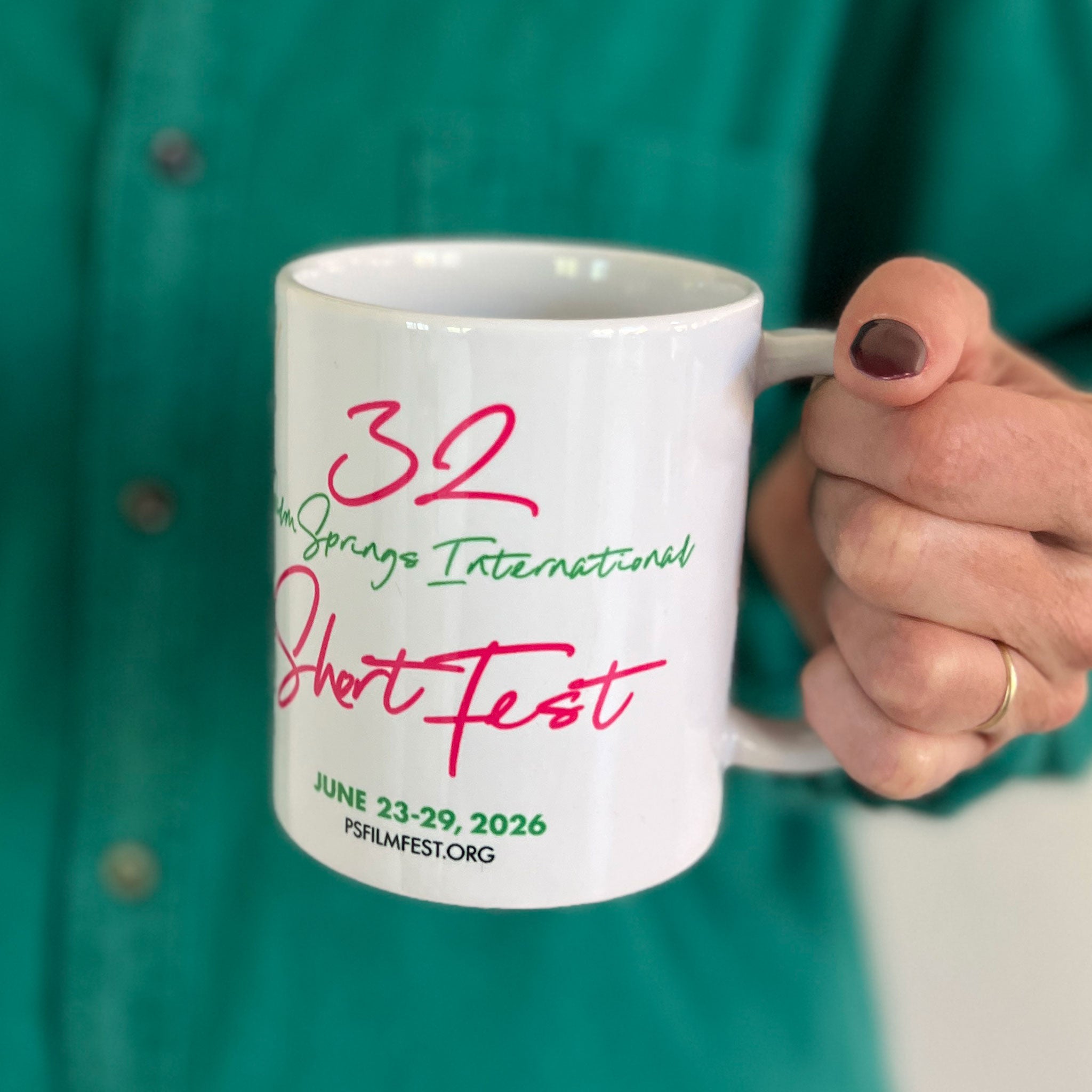 White mug with '32 Palm Springs International ShortFest' text held by a person in a green shirt.