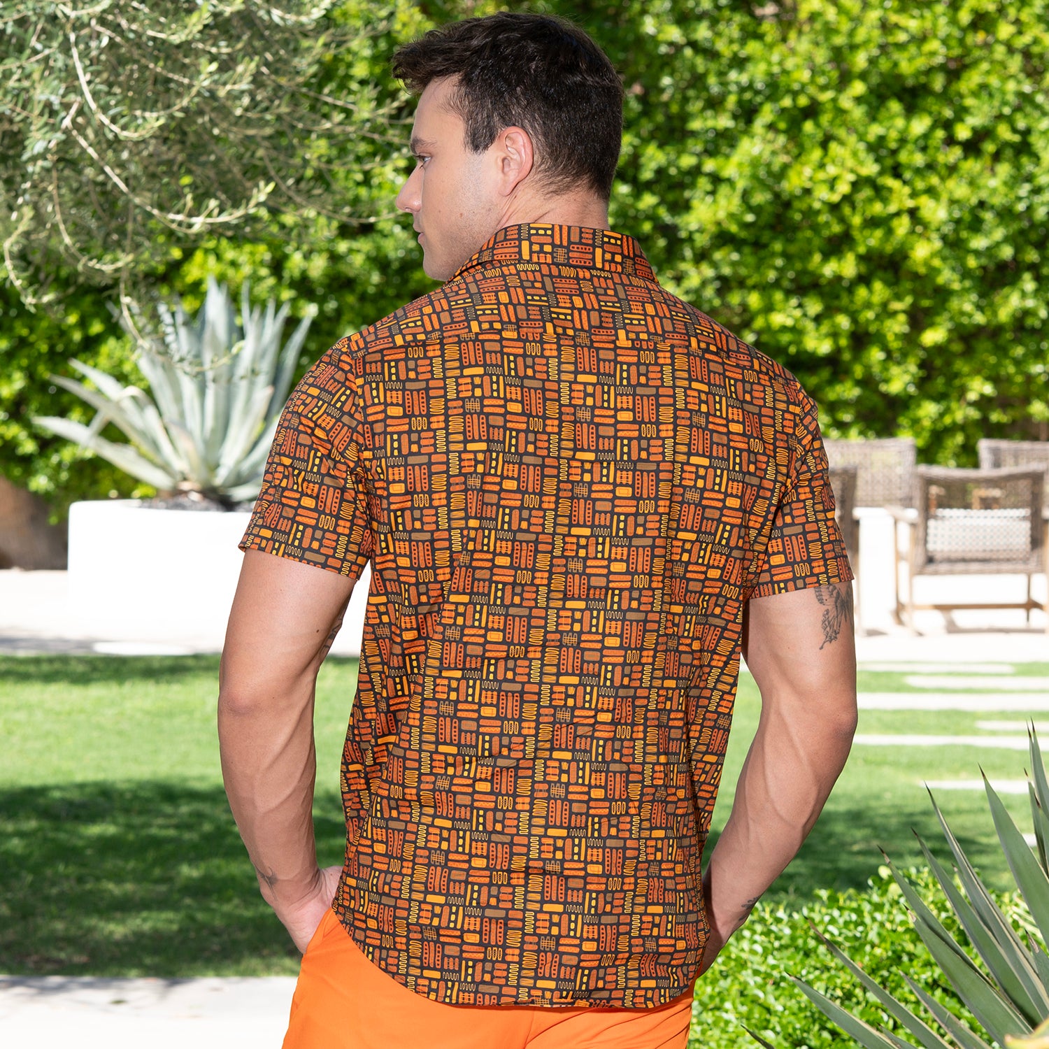 A man viewed from behind wearing a short-sleeve button-up shirt with an orange, brown, and black geometric pattern, set against a lush green outdoor background.