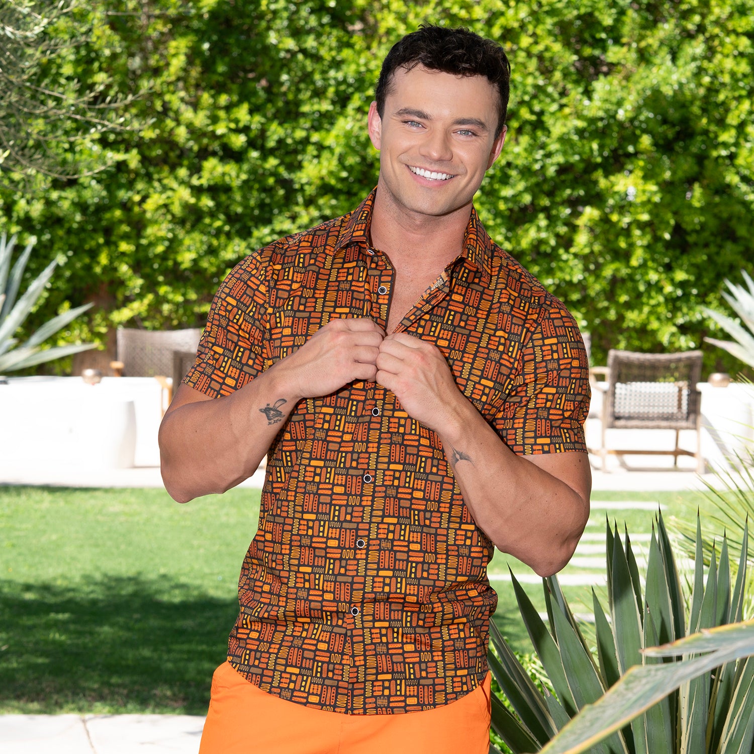 A smiling man wearing a short-sleeve button-up shirt with an orange, brown, and black geometric pattern, paired with bright orange pants, standing outdoors in a garden setting.