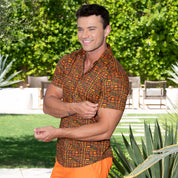 A smiling man standing outdoors in a garden setting, wearing a short-sleeve button-up shirt with an orange, brown, and black geometric pattern and bright orange pants.