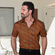 A man wearing a short-sleeve button-up shirt with a dense geometric block pattern in orange, yellow, and black, paired with black pants, standing in a brightly lit modern interior.