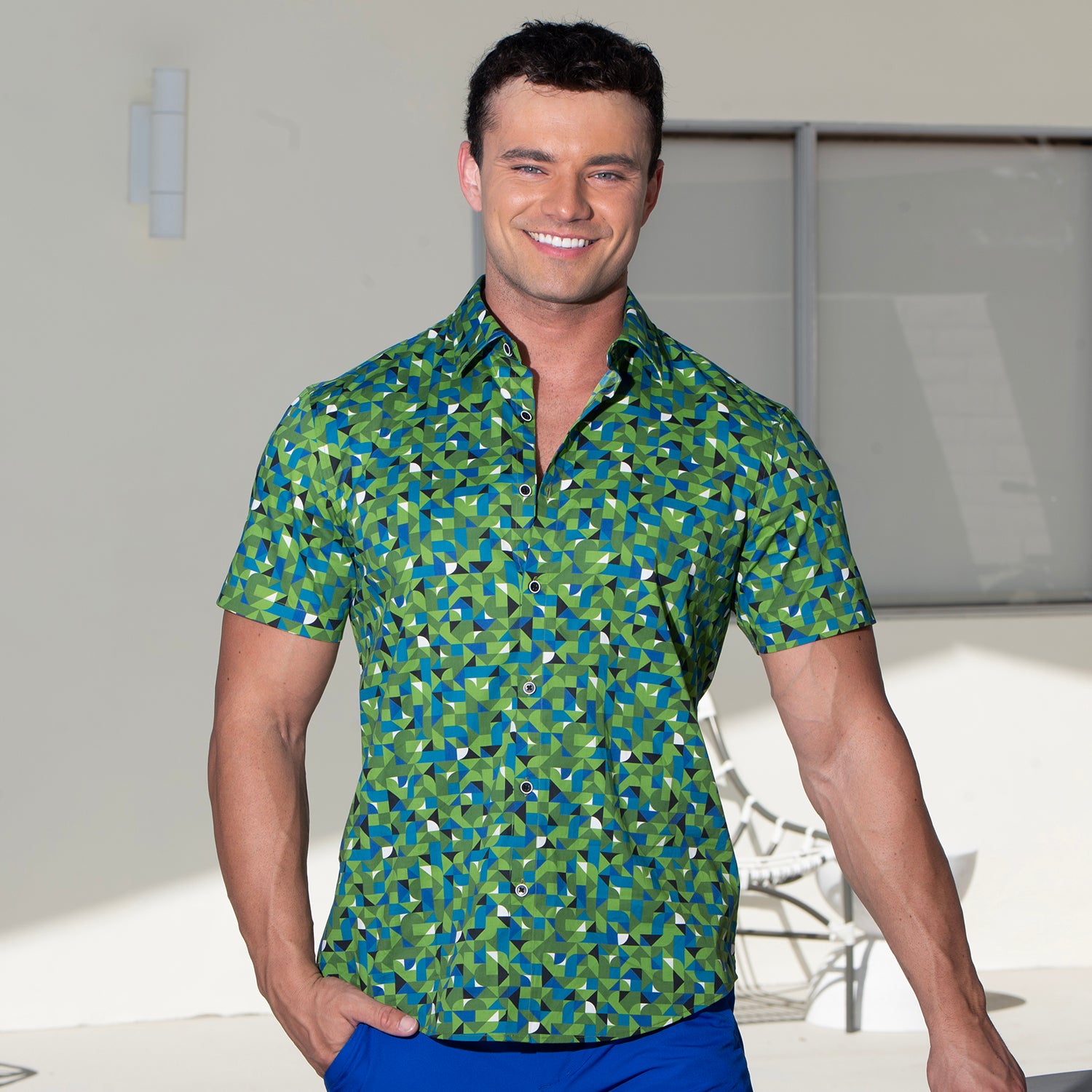 A smiling man wearing a short-sleeve button-up shirt with a green, blue, and white geometric pattern, paired with blue pants, standing against a light-colored wall.
