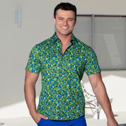 A smiling man wearing a short-sleeve button-up shirt with a green, blue, and white geometric pattern, paired with blue pants, standing against a light-colored wall.