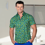 A smiling man wearing a short-sleeve button-up shirt with a green, blue, and white geometric pattern, paired with bright blue pants, standing in a brightly lit modern interior.