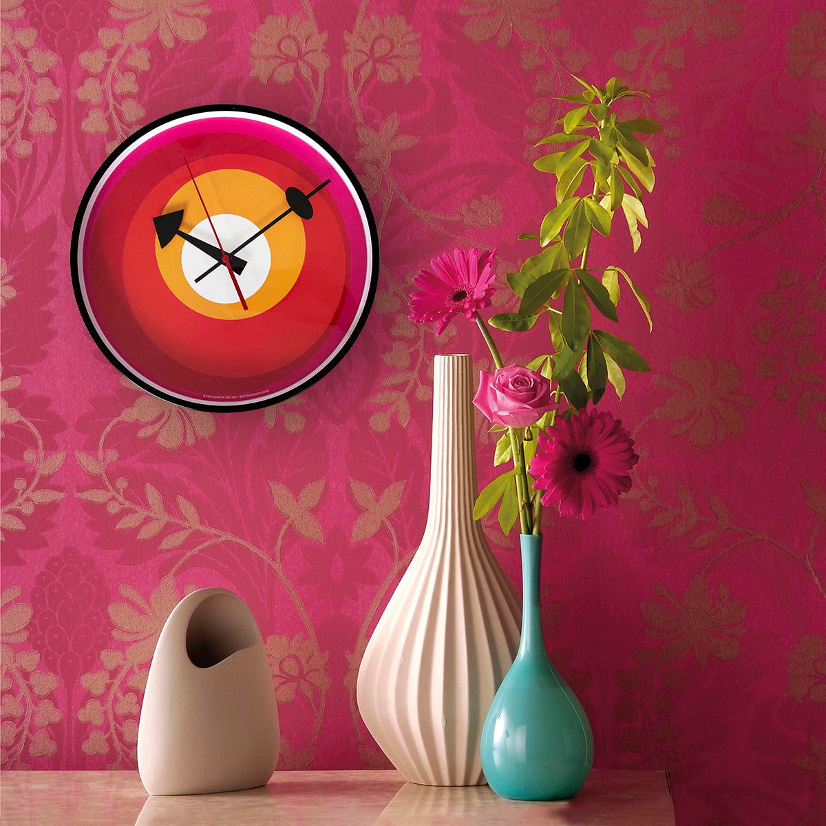 mod wall clock with no numbers but just red, yellow, pink and white circles hanging on a floral pink wall with vases and flowers
