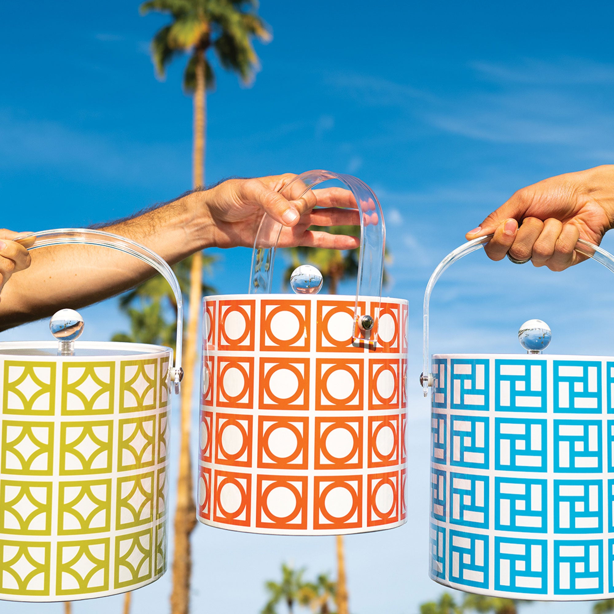 Three patterned ice buckets held by hands against a blue sky with palm trees.