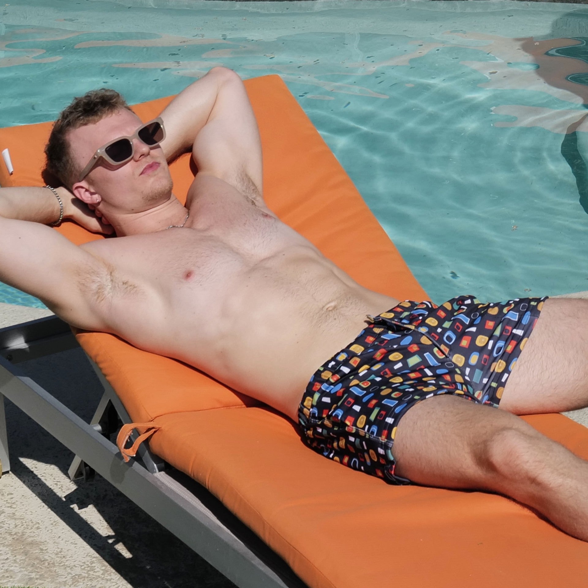 A shirtless man wearing patterned swim trunks and sunglasses relaxes on an orange pool lounge chair by a swimming pool.