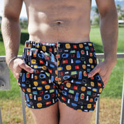 A man wearing black swim trunks with a colorful geometric pattern of red, yellow, blue, and green shapes, standing outdoors with a grassy background.