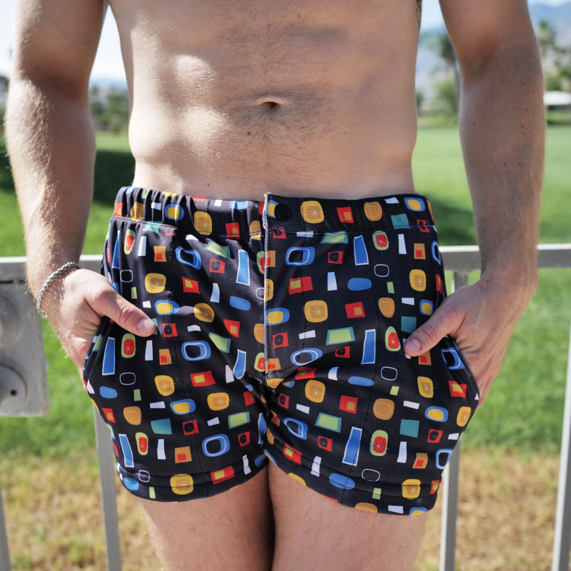 A man wearing black swim trunks with a colorful geometric pattern of red, yellow, blue, and green shapes, standing outdoors with a grassy background.