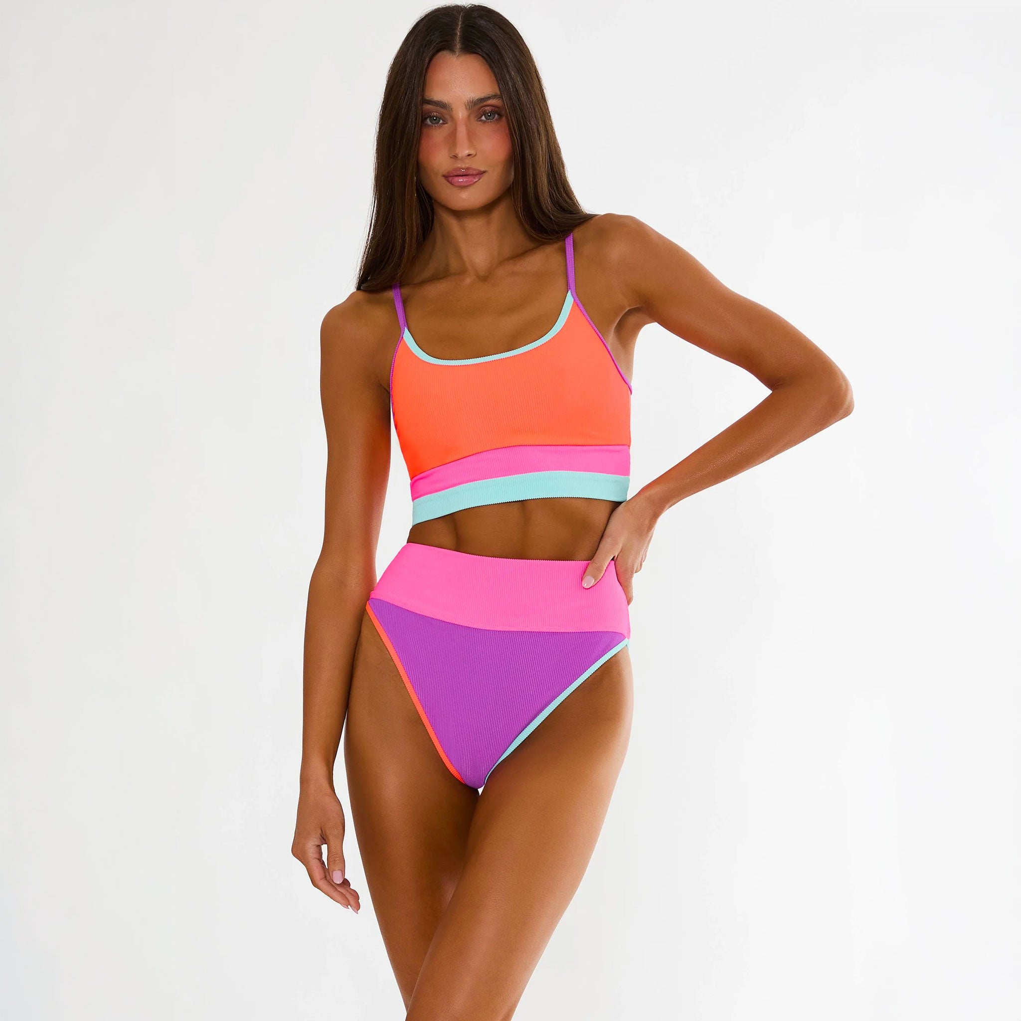 A woman modeling a bright color-blocked two-piece swimsuit with a coral orange top, pink and light blue bands, and purple high-waisted bottoms, set against a plain white studio background.