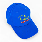 A royal blue baseball cap featuring embroidered 'Palm Springs' text on the front panel, with 'Palm' in yellow script and 'Springs' in red block letters, shown on a clean white background.