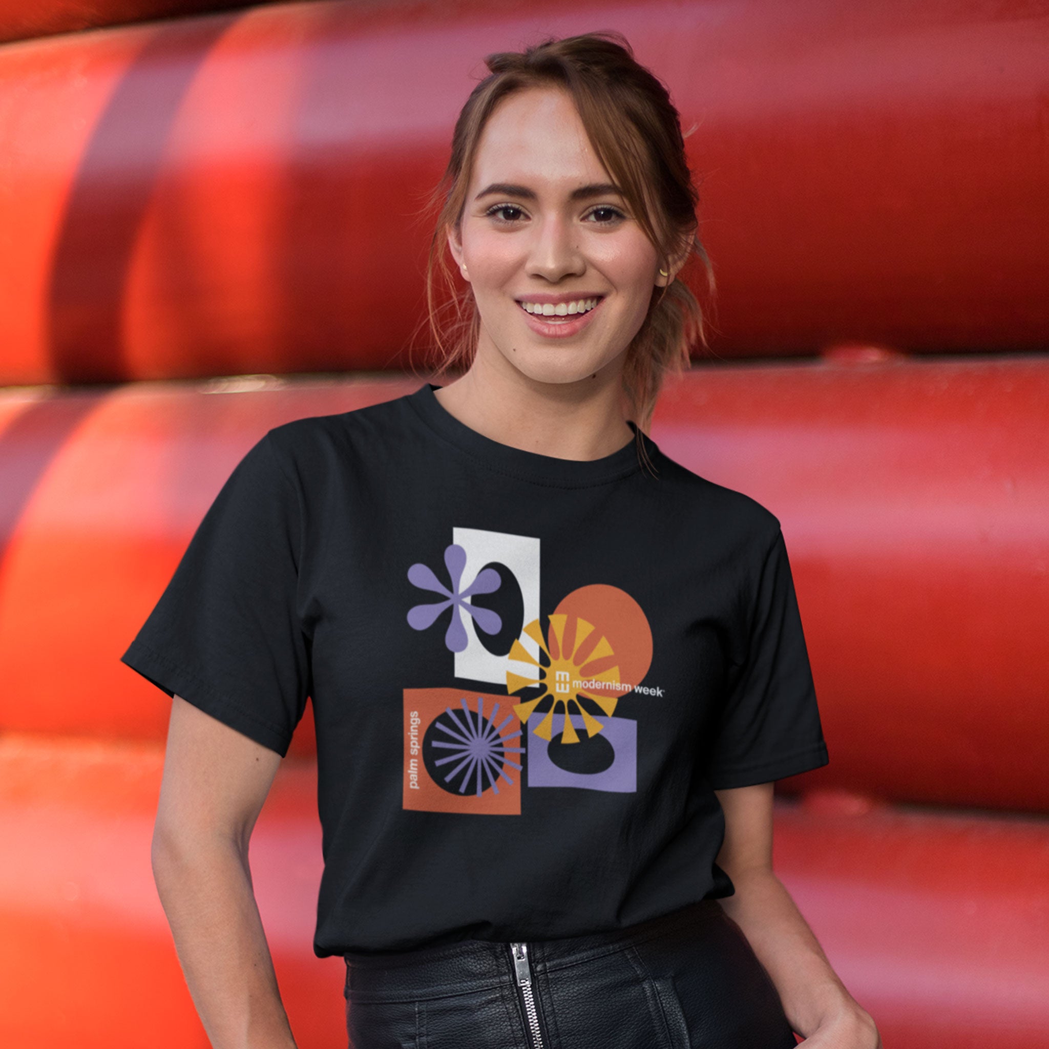 A woman wearing a black graphic t-shirt with a colorful mid-century modern geometric print, standing in front of a red industrial background.