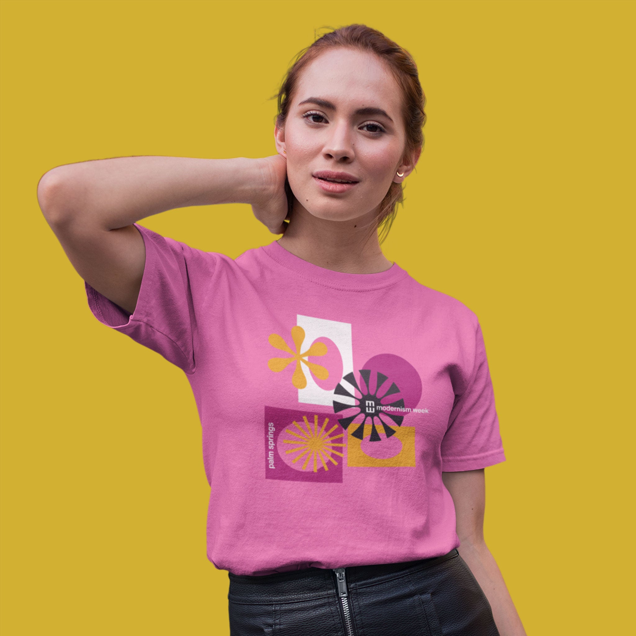A woman modeling a pink graphic t-shirt with a retro 1970s-style abstract design in yellow, purple, and white, set against a solid yellow background.