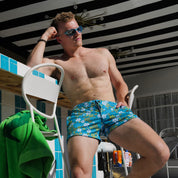 A shirtless man wearing blue floral swim trunks and sunglasses leans against a white chair in a poolside setting with a striped ceiling overhead.