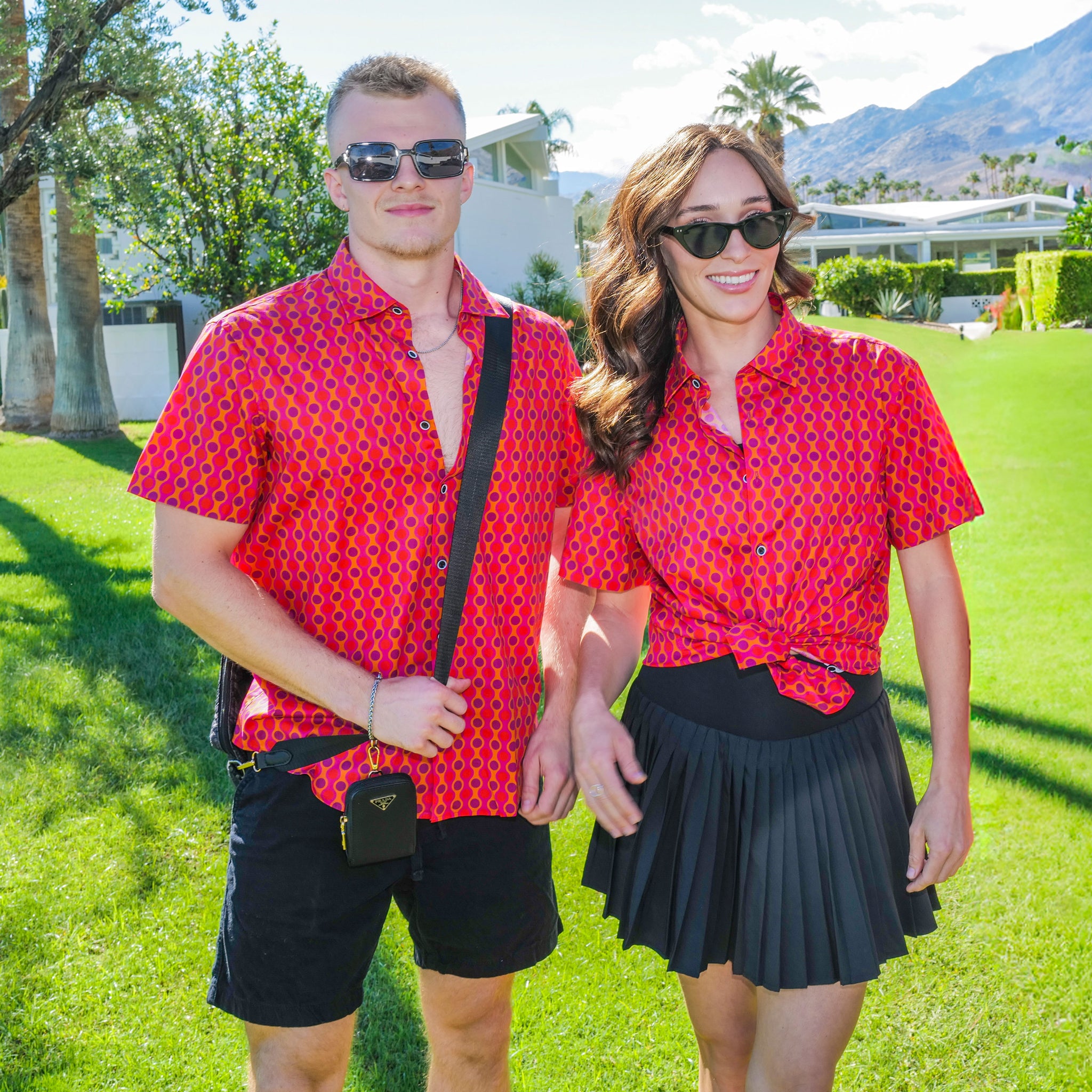Two people wearing matching red patterned shirts on a grassy outdoor setting with palm trees and mountains in the background.