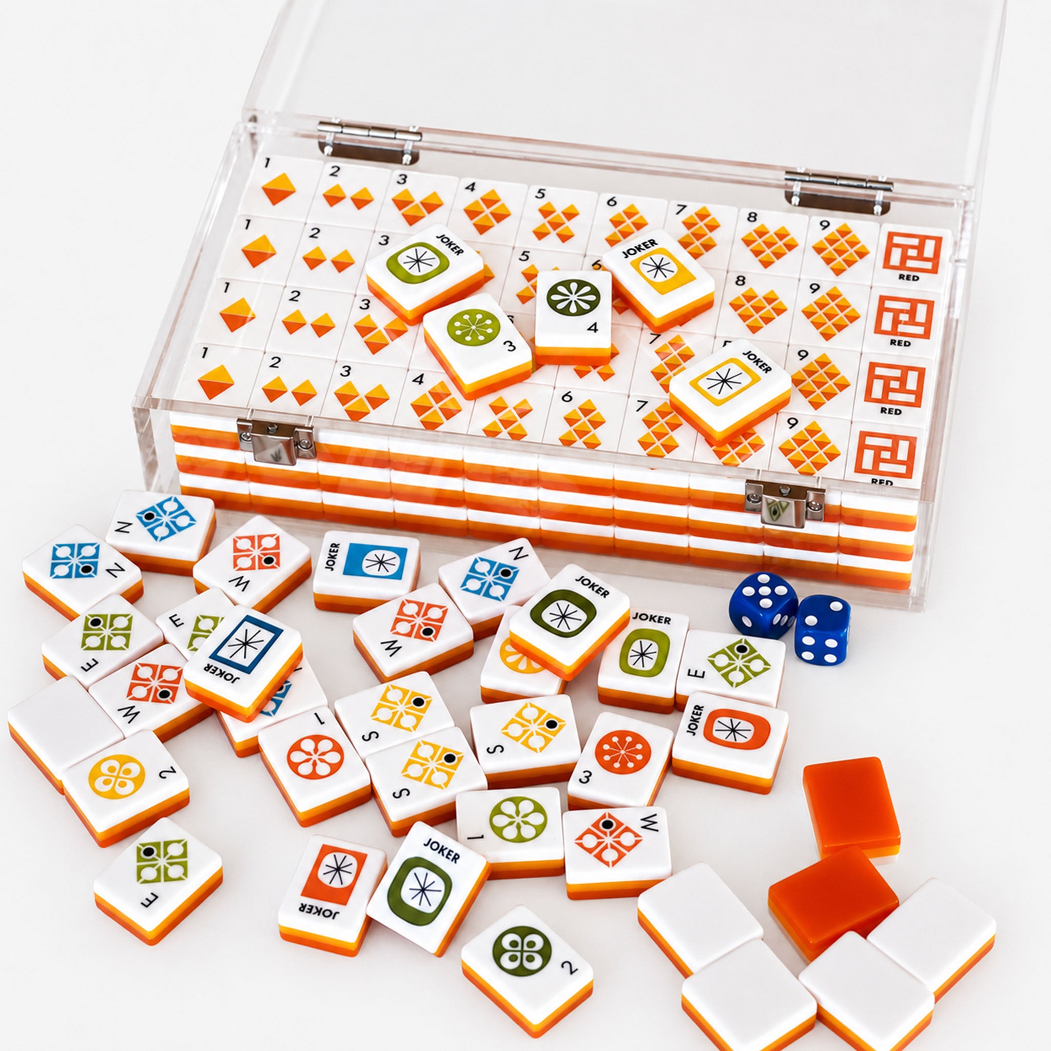A clear plastic case filled with Mahjong tiles, with several loose tiles and two blue dice scattered in front on a white background.