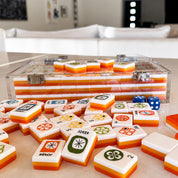 A set of mahjong tiles with a clear acrylic case, shown with several tiles scattered in the foreground on a tabletop. The tiles are primarily white with orange borders and multicolored symbols, with blue dice visible.