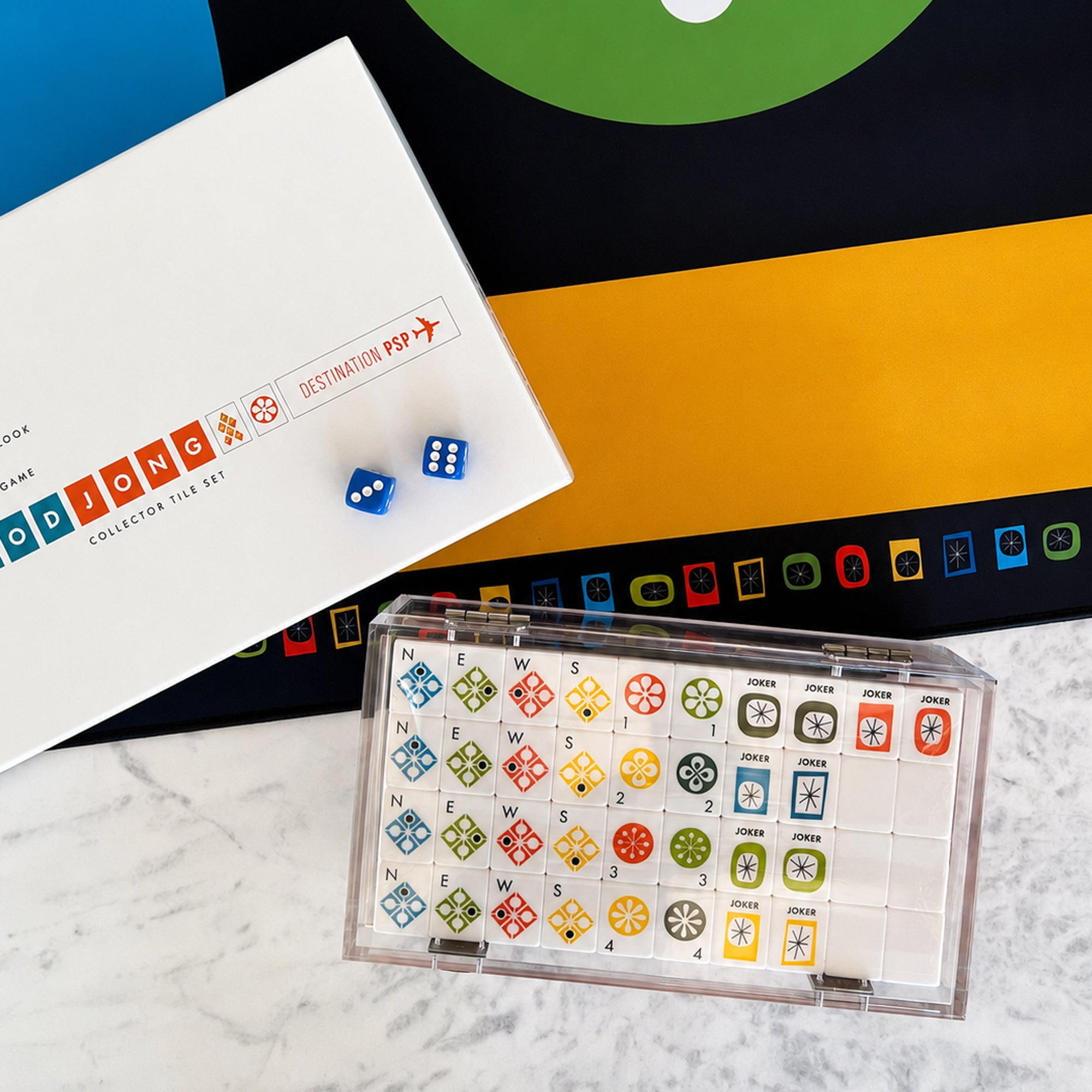A board game set displayed on a marble surface, including a white collector's box labeled 'Destination Pop!', a clear plastic game board with colorful character tiles, and two blue dice.