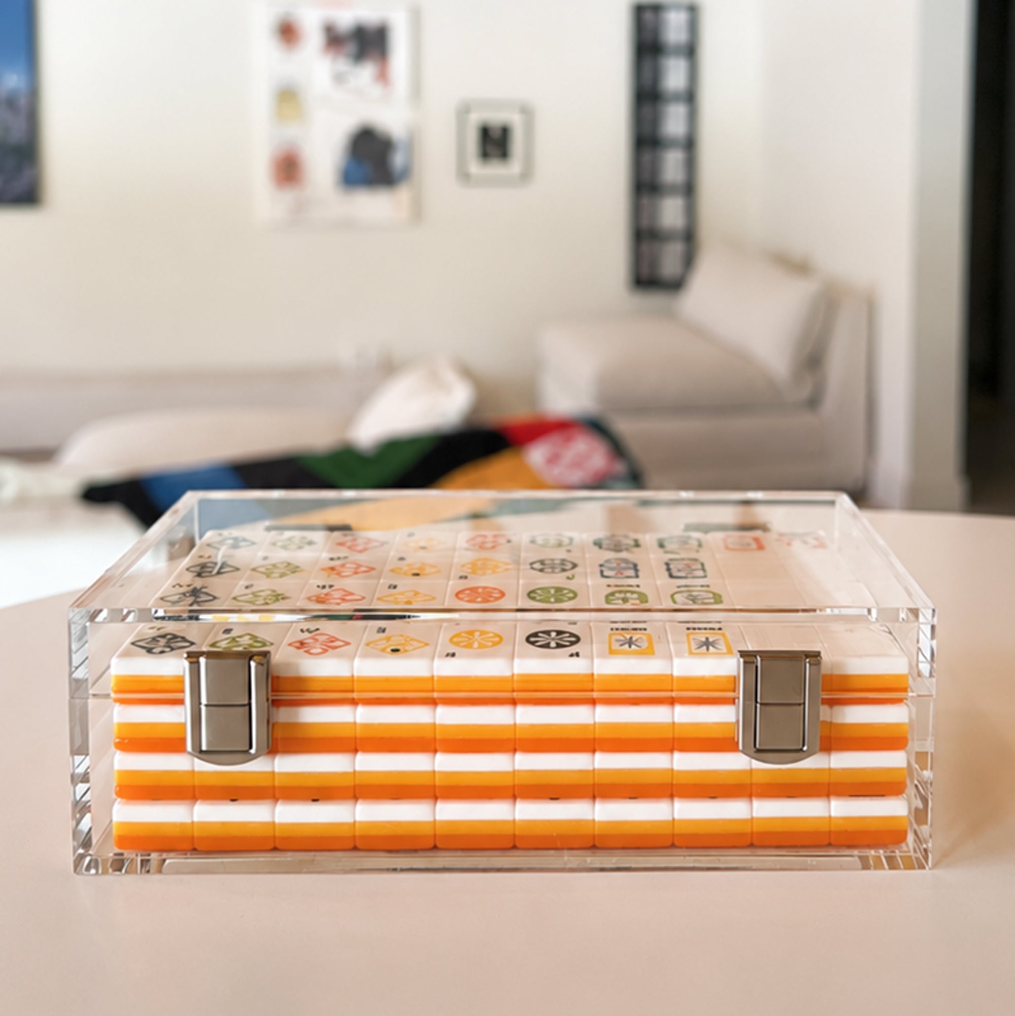 A clear acrylic mahjong case with orange and white striped backing and a transparent top displaying game tiles, set on a light-colored table with a blurred living room background.