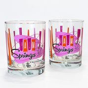 Two clear glass rocks tumblers with a retro 'Palm Springs California' design featuring a pink and purple abstract shape, orange and red vertical stripes, and black script text, shown on a white background.