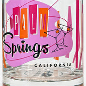A clear glass tumbler featuring a colorful retro-style 'Palm Springs California' graphic with pink, orange, and black lettering and line art.