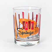 A clear glass rocks tumbler featuring a retro 'Palm Springs California' design with orange and red geometric stripes and black script text, shown on a light gray background.