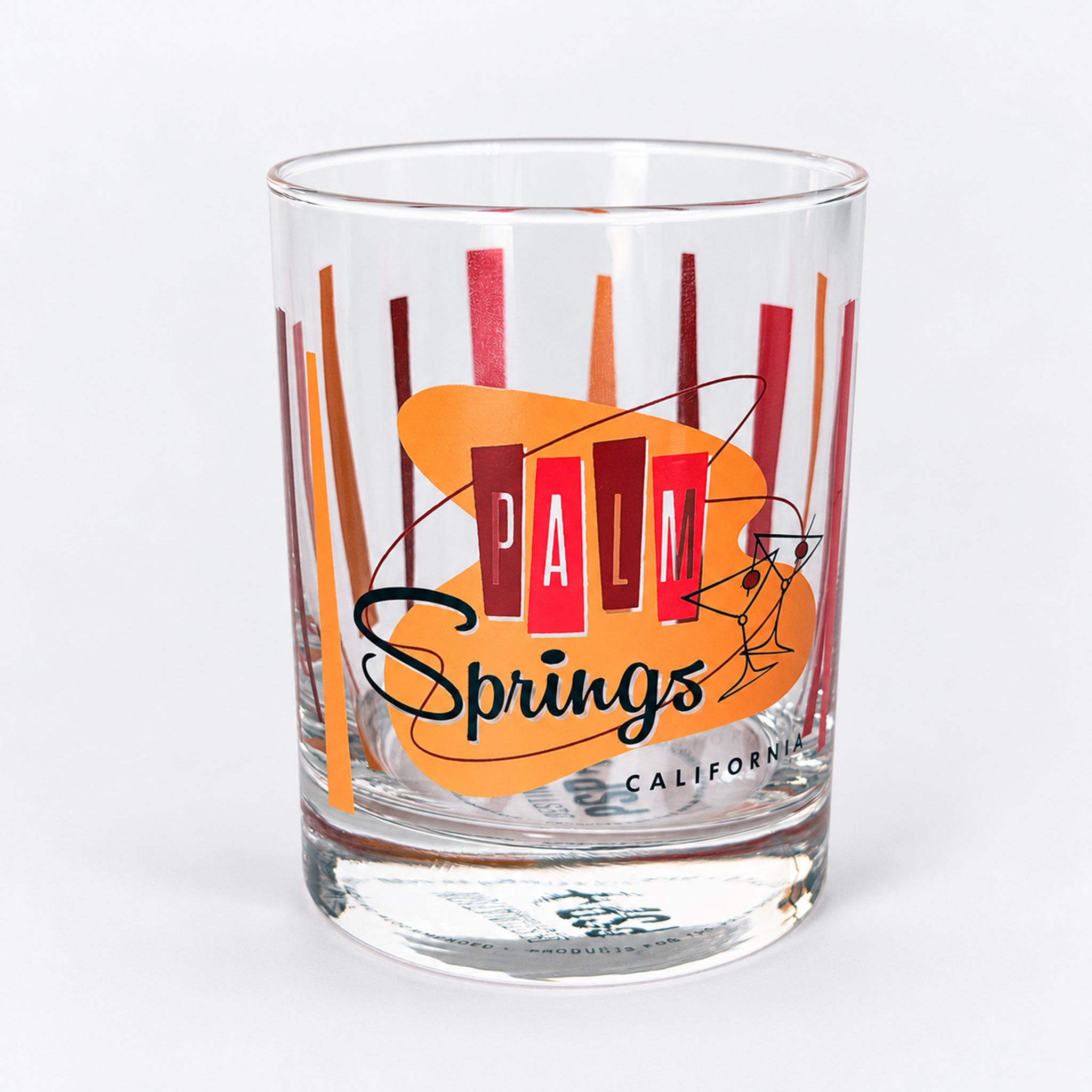 A clear glass rocks tumbler featuring a retro 'Palm Springs California' design with orange and red geometric stripes and black script text, shown on a light gray background.
