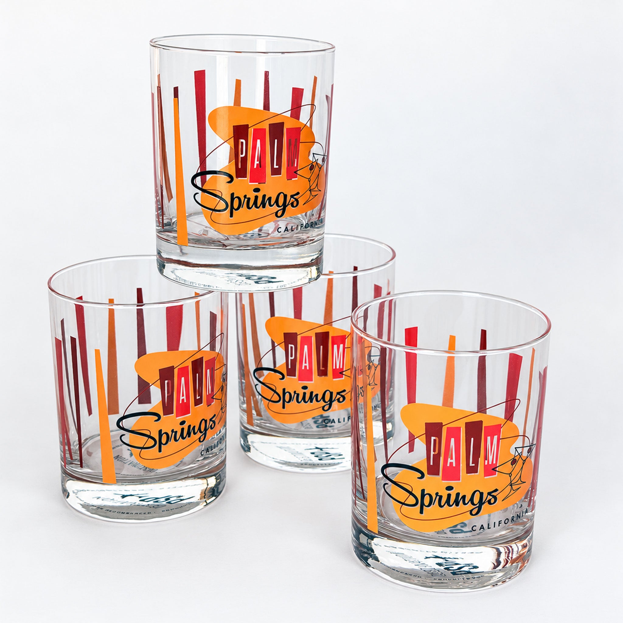 Four clear glass tumblers with a vintage-style 'Palm Springs California' print, featuring orange and red abstract shapes and black script, arranged on a white background.