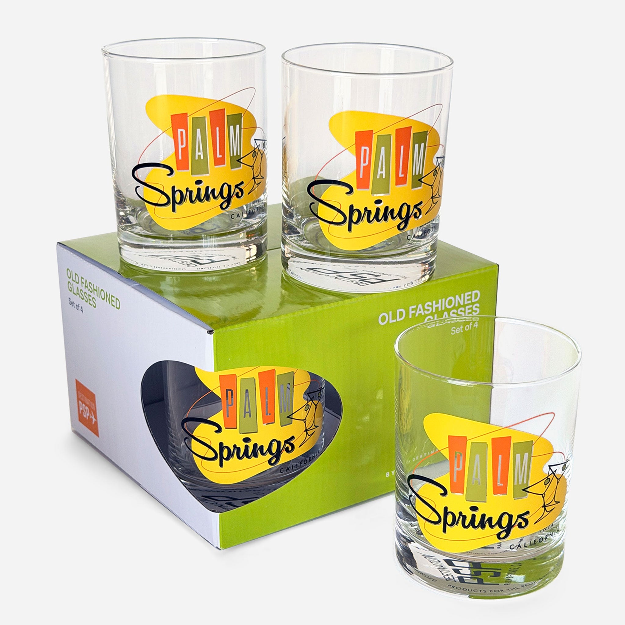 Three clear Palm Springs Old Fashioned glasses shown with one in front and two stacked on top of their green and white retail box. The glasses have a yellow retro sun graphic with the words 'Palm Springs' in multicolored block letters and black script.