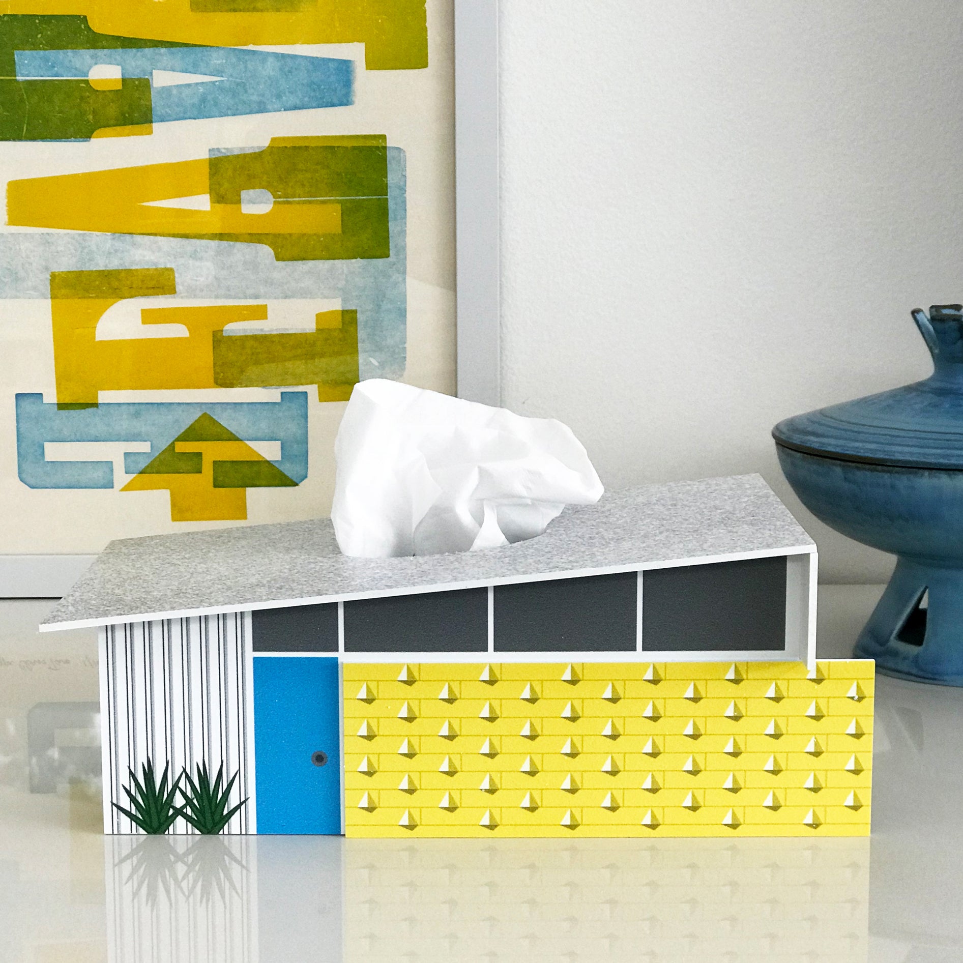 A midcentury modern tissue box on top of a sleek tabletop with vibrant art in the background.