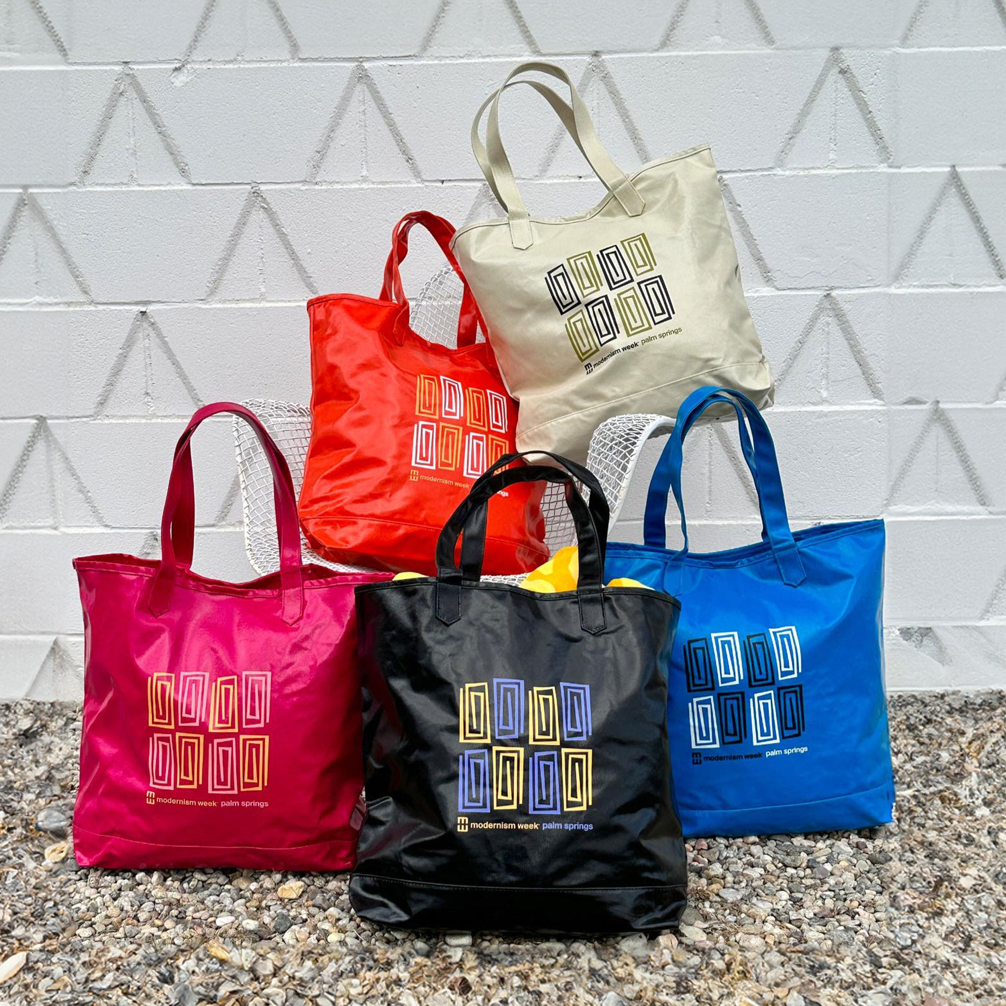 Five colorful reusable shopping tote bags stacked against a midcentury modern brick wall.