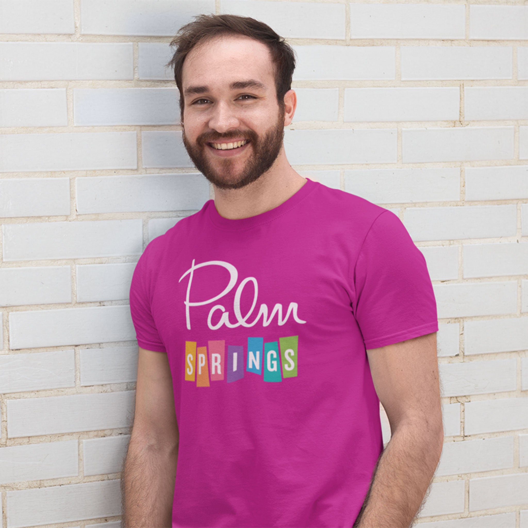 A man smiling and laughing, wearing a bright pink t-shirt with 'Palm Springs' printed on the front, set against a solid yellow background.