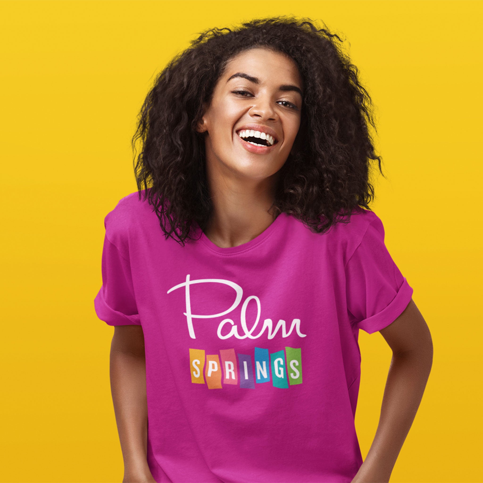 A woman with curly dark hair smiling and laughing, wearing a bright pink t-shirt with 'Palm Springs' printed on the front, set against a solid yellow background.