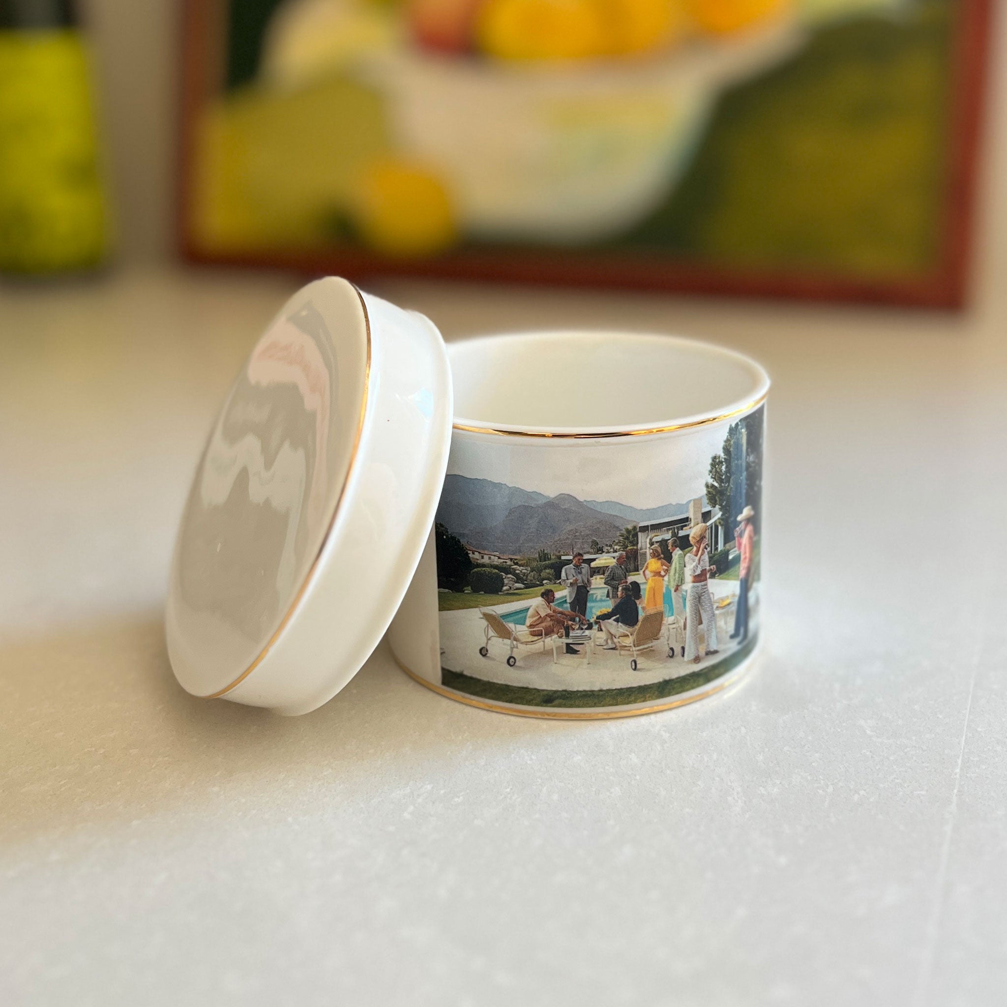 A small white ceramic keepsake box with a lid, featuring a printed scenic landscape band around the body and gold trim, set on a light surface with a blurred background.