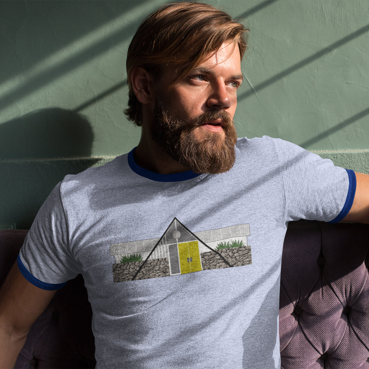 Man with beard sitting on couch looking out the window wearing ringer blue T-shirt.